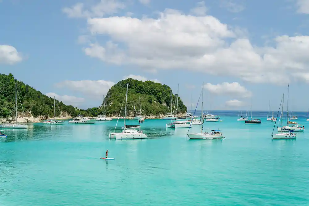 a green islet ringed with anchored yachts and a paddleboarder crossing the bay