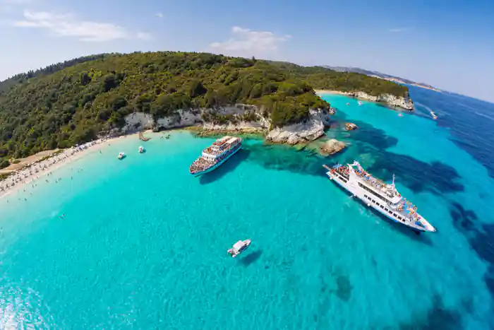 two cruise boats anchored over turquoise shallows below a wooded Antipaxos headland